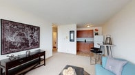 This single-floor unit features an open-concept design that seamlessly connects the living area and kitchen, creating a cohesive and functional layout. The living area is finished with plush wall-to-wall carpeting, providing a comfortable and inviting atmosphere. White walls throughout keep the space feeling bright and clean, complemented by flush-mount ceiling lighting that ensures consistent illumination. A decorative framed art piece anchors one wall of the living area, adding visual interest.

The kitchen is partially separated by a half-wall partition, offering a degree of definition between the two spaces while maintaining an open feel. It features warm medium-toned wood cabinetry with dark countertops, a white refrigerator, and tile flooring that contrasts with the carpeted living area. A compact breakfast bar with black stools provides casual seating. A hallway visible from the living area leads to additional rooms, suggesting a practical and efficient floor plan throughout the unit.