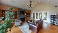 This thoughtfully designed single-floor community space exudes warmth and comfort through a blend of classic architectural details and inviting furnishings. The centerpiece of the main lounge is a dramatic floor-to-ceiling stacked stone fireplace topped with a mounted television, flanked by rich wood built-in shelving and cabinetry. Soaring vaulted ceilings amplify the sense of openness, while an elegant wrought-iron chandelier adds a refined touch overhead. Rich hardwood flooring flows throughout, grounding the space with warmth and continuity. Generous natural light pours in through large arched windows and multiple sets of white-framed French doors, offering views of an outdoor pool area surrounded by lush greenery. The lounge is furnished with plush leather sofas, upholstered armchairs, and a media and book display rack, creating a relaxed yet functional gathering environment. A pass-through opening connects the main lounge to a well-appointed kitchenette, which features warm maple cabinetry, dark countertops, stainless steel appliances, and a window overlooking the pool. The space is ideal for community gatherings, social events, or quiet relaxation, combining practical amenities with an inviting, residential ambiance throughout.
