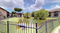 This single-story residence is situated within a well-established community that offers a notably serene outdoor environment. The grounds feature a decorative pond complete with an active fountain, framed by sweeping willow trees and mature shade trees. Lush green lawns extend throughout the property, and a wrought iron perimeter fence adds a classic, finished aesthetic to the exterior setting.

Inside, the interior is defined by a clean, neutral palette with warm cream-toned walls and dark hardwood-style flooring that grounds each space with a sense of depth and warmth. Recessed lighting provides even illumination throughout. The dining area serves as a standout interior feature, centered around a striking round green marble table paired with woven rattan chairs atop a natural fiber rug. A large-scale framed artwork anchors one wall, contributing an artistic, gallery-like quality to the room.

Distinctive architectural details include a sliding barn door and a set of rustic double doors that connect interior spaces, adding character and visual interest. Framed wall art displayed in the hallway further reinforces the curated, design-forward sensibility that runs throughout this thoughtfully appointed single-floor residence.