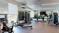 The Vela Fitness Center is a sleek, single-floor facility designed to support a comprehensive workout experience. The space features a clean, modern aesthetic with white walls, linear pendant lighting, and large windows fitted with roller shades that invite abundant natural light while maintaining a bright, open atmosphere. Full-length mirrors line multiple walls throughout the space, visually expanding the room and allowing users to monitor their form during exercise.

The fitness center is thoughtfully organized into distinct workout zones. A dedicated cardio area houses a collection of Matrix ellipticals, stationary bikes, and rowing machines, positioned to take advantage of the natural light from the expansive windows. The strength training section includes a multi-functional cable machine system, a full rack of dumbbells ranging across multiple weights, and a variety of resistance and weight machines to accommodate both upper and lower body training. A large flat-screen television is mounted on the mirrored wall, providing entertainment during workouts.

The flooring throughout consists of durable rubber gym matting, ensuring safety and comfort. Entry to the fitness center is through a stylish glass-and-dark-metal framed door. The overall ambiance is professional and contemporary, reflecting the high standards of The Vela community in Tracy, California.