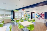 Located at 12121 Greenspoint Drive in Houston, TX, this lively community room is a thoughtfully designed, single-floor space that seamlessly blends functionality with artistic energy. The open layout accommodates multiple white square tables paired with bright lime-green chairs, offering flexible seating arrangements ideal for gatherings, activities, or collaborative use.

The interior design is defined by bold, expressive watercolor splatter murals adorning multiple walls, injecting a dynamic burst of color throughout the space. Complementing these murals are striking navy blue accent walls and a series of colorful circular pendant light fixtures — in green, red, and blue — suspended from the white ceiling, adding both illumination and visual interest overhead.

Wood-look flooring runs throughout, providing a warm, grounded contrast to the vibrant palette above. Multiple windows with roller shades allow ample natural light to filter in, offering views of the surrounding outdoor landscape.

A dedicated kitchenette area features bold navy blue cabinetry, a multicolored mosaic tile backsplash, a countertop with a sink, and a beverage cooler beneath — making the space well-equipped for hosting events. Recessed lighting supplements the pendant fixtures, ensuring the room is well-lit throughout. The overall ambiance is energetic, welcoming, and creatively stimulating.
