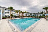 Located at 6136 Blanding Boulevard in Jacksonville, Florida, this outdoor amenity space is designed as a resort-style retreat that centers around a generously sized swimming pool with crystal-clear blue water. The pool deck is finished in light-toned pavers, providing a clean and polished aesthetic that complements the surrounding contemporary architecture.

The deck is well-appointed with a variety of seating options, including rows of cushioned chaise lounges and coordinating side tables positioned directly along the pool's edge for convenient access. Vibrant blue patio umbrellas offer shaded relief throughout the terrace, while additional lounge groupings with soft seating and pergola-style canopies create defined social areas for relaxation and gathering.

Mature palm trees are thoughtfully integrated throughout the deck, lending a tropical, resort-like ambiance to the space. The pool is enclosed by a black iron perimeter fence, providing both security and an unobstructed sightline to the broader community.

The adjacent one-story clubhouse features gray lap siding, white trim, and a covered overhang with ceiling fans, allowing for comfortable outdoor use in warm weather. Large glass sliding doors connect the interior amenity spaces directly to the pool deck, creating a seamless indoor-outdoor flow. The multi-story residential buildings surrounding the pool area feature balconies that overlook this central outdoor gathering space.