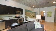 This single-floor studio presents an open-concept layout that efficiently combines living, sleeping, dining, and cooking areas within one cohesive space. The exposed concrete ceiling serves as a defining architectural element, lending an industrial character that contrasts with the clean white walls and contemporary finishes throughout.

The kitchen is positioned along one wall and features dark espresso cabinetry, stainless steel appliances including a range and over-the-range microwave, and a light-toned countertop. A dark-finished breakfast bar extends from the kitchen, offering a casual dining or workspace surface.

The living area centers around a grey upholstered sofa accented with green and white patterned throw pillows, creating a comfortable lounge space. Flowing seamlessly from the living area, the sleeping zone is anchored by a full-sized bed with a tall upholstered headboard set against a teal accent wall, adding a pop of color to the neutral palette.

Recessed lighting supplements the space, while a table lamp beside the bed contributes warm ambient light. A partially visible bathroom with dark cabinetry and hardwood-style flooring rounds out the unit's functional offerings.