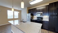 This single-floor residence showcases a well-appointed kitchen designed for both functionality and aesthetic appeal. At the heart of the space is a generous island topped with light-toned granite countertops, featuring an undermount sink with a contemporary arc faucet — an ideal workspace for meal preparation and casual dining. The kitchen is anchored by rich, dark espresso-finished cabinetry that extends to the upper walls, providing ample storage while contributing a bold, modern contrast against the warm neutral wall tones. Stainless steel appliances, including a refrigerator and over-the-range microwave, complement the sleek design, while a black electric range adds a striking accent. A subway tile backsplash lines the cooking area, adding texture and visual interest. Overhead, pendant lights hang above the island, providing focused task lighting alongside flush-mounted ceiling fixtures. Natural light filters into the kitchen through blinds-fitted windows, brightening the space and enhancing its open, airy feel. Hardwood-style flooring grounds the overall design, tying together the contemporary elements throughout the kitchen.