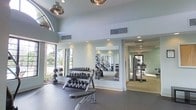 This fitness center is designed to support a comprehensive workout experience across a single thoughtfully organized floor. The space is anchored by soaring ceilings that lend an open, airy quality, complemented by large arched windows that flood the free weights area with natural light and views of the lush greenery outside. Recessed lighting and stylish pendant fixtures provide ample illumination throughout. The free weights zone features a full rack of dumbbells alongside medicine balls and a workout bench, with a large wall-mounted mirror that enhances the sense of space and allows for proper form checks during exercise. Durable dark rubber flooring runs through the primary workout area, offering both safety and performance underfoot. Adjacent to the free weights section, a separate room houses cardio and cable machines, finished with lighter flooring and continued recessed lighting for a clean, functional environment. Neutral tones of soft gray and white on the walls contribute to a calm, focused atmosphere throughout. Ventilation is well-integrated with visible ceiling ducts ensuring comfort during intense workouts. The overall layout is efficient and practical, making this fitness center a well-rounded facility suited for a variety of training routines.