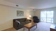 This single-floor residence features a welcoming living room designed for comfort and functionality. The space is finished with wall-to-wall carpeting that adds warmth underfoot, while neutral-toned walls create a clean, versatile backdrop suitable for a variety of décor styles. A ceiling-mounted HVAC vent ensures year-round climate control throughout the room.

The living area is well-proportioned, offering enough room to accommodate a full-sized sofa and a leather recliner chair, arranged around a classic wooden coffee table. A small side table with a table lamp provides ambient lighting, complementing the natural light that enters through a large sliding glass door fitted with vertical blinds. The sliding door appears to lead to a private outdoor area, allowing for both ventilation and exterior access.

Wall space is utilized for decorative purposes, with room for framed artwork that adds a personalized touch to the interior. The overall ambiance of the living space is casual and inviting, making it a comfortable everyday environment. The straightforward layout maximizes usable floor space while maintaining an open, uncluttered feel throughout.
