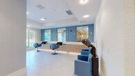 Located at 1231 Arya Way in Ocoee, Florida, this single-floor professional salon is thoughtfully designed with a clean, contemporary aesthetic. The main styling area is the centerpiece of the space, featuring multiple styling stations lined along a light wood cabinetry wall accented by blue tile paneling and large mirrors. Blue upholstered styling chairs complement the cohesive color palette, while recessed lighting keeps the environment bright and welcoming. Built-in shampoo bowls with sleek black basins are integrated into the cabinetry, providing a seamless and functional layout. Dedicated hair dryer chairs are positioned along the opposite wall, offering a comfortable and efficient drying area. Large windows flood the styling area with natural light and provide views of the surrounding outdoor greenery, enhancing the open and airy ambiance. A separate nail care room offers a more private setting, equipped with a pedicure chair and a manicure table with coordinating blue seating. Light tile flooring throughout the space maintains a bright, polished appearance. A wide, well-lit corridor with reflective mirrored accents connects the different service areas, contributing to the sense of spaciousness. The overall design balances functionality and modern style, creating a comfortable and professional environment for a full range of salon services.