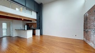 This two-story loft-style space masterfully combines industrial character with modern finishes throughout. The main level immediately impresses with soaring double-height ceilings and warm hardwood flooring that extends across the expansive open living area. Distinctive exposed steel columns with cross-bracing serve as architectural focal points, lending an authentic industrial aesthetic. A staircase with a sleek metal railing connects the two floors, with the upper level featuring an open railing overlooking the main living space below.

The kitchen is well-appointed with warm wood cabinetry, dark granite countertops, and stainless steel appliances, including a full-size refrigerator. A breakfast bar provides additional prep and seating space. Wood-paneled ceilings and track lighting add warmth and dimension to the kitchen area.

One room features dramatic ceiling height with clerestory windows and a skylight that flood the space with natural light, alongside a striking exposed brick accent wall that reinforces the building's historic character.

The upper level offers carpeted bedroom spaces with natural light from multiple windows, a walk-in closet, and an en-suite bathroom complete with a granite-topped vanity, tub-shower combination, and clean white tile finishes. An additional galley-style kitchen with hardwood floors and tall windows rounds out the versatile layout.