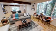This single-floor space showcases a cohesive mid-century modern aesthetic carried throughout its open layout. The main living area is anchored by a substantial grey sofa accented with bold houndstooth and textured throw pillows, complemented by a warm walnut-toned coffee table resting atop a grey area rug. A distinctive exposed dark wood ceiling beam adds architectural character and visual depth to the space.

Natural light flows generously through large windows on multiple walls, offering views of surrounding mature trees and greenery, creating a seamless indoor-outdoor connection. The warm hardwood flooring runs throughout, unifying the living and dining areas. A compact round dining table with mid-century style tapered legs sits adjacent to the seating area, paired with an eclectic patchwork accent chair that adds a playful pop of color.

The space transitions into a secondary seating area, visible in the background, featuring curved barrel chairs in muted tones alongside a wooden dresser, suggesting a versatile multi-use layout. Recessed lighting supplements the abundant natural light. Wall art featuring a retro travel poster adds a personalized, curated touch to the overall warm and inviting ambiance.