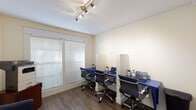 Located at 4711 S Himes Ave in Tampa, Florida, this single-floor professional workspace offers a well-organized and inviting environment designed to support productivity and client comfort. The layout flows from a welcoming entryway — featuring warm wood-look flooring, white paneled French doors with glass panes, and tasteful wall art — into distinct functional areas throughout the space.

The primary workstation room is equipped with a wall-mounted blue desk running the length of the room, divided into individual stations by clear privacy panels. Each station is paired with a modern ergonomic chair, providing a clean and focused environment for multiple users. Track lighting overhead ensures consistent, well-lit conditions, while large windows fitted with horizontal blinds allow natural light to fill the room.

A relaxed reception or waiting area features a plush blue accent chair positioned beneath a large decorative wall clock with a rustic wood face and Roman numerals, adding a warm, stylistic focal point to the space. Lush greenery and a wood-toned storage unit complete the area. Louvered bi-fold closet doors provide additional storage access within the space.

Throughout, the consistent use of wood-look flooring, neutral cream walls, and white trim creates a cohesive, professional aesthetic that is both functional and visually appealing.
