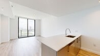 Located on the 19th floor in Miami's vibrant Edgewater neighborhood, this one-bedroom, one-bathroom residence offers a clean, contemporary aesthetic throughout. Light wood-tone flooring flows seamlessly across the open-plan living and kitchen area, creating a bright and cohesive feel. Floor-to-ceiling windows with black-framed sliding glass doors flood the living space with natural light while framing sweeping views of the Miami skyline and surrounding greenery. A private balcony extends the living area outdoors, ideal for enjoying the city panorama.

The kitchen is thoughtfully designed with a peninsula island featuring a white quartz countertop and an integrated sink, complemented by gold-toned cabinetry, white upper cabinets, and a full suite of stainless steel appliances including a refrigerator, gas range, and built-in microwave. Recessed lighting illuminates the workspace efficiently.

The spacious bedroom benefits from its own set of floor-to-ceiling windows, a ceiling fan, and a built-in closet, offering both comfort and ample storage. The bathroom is finished in a modern style with a white quartz vanity, sleek cabinetry with gold hardware, and large-format tile flooring. The unit's crisp white walls and minimalist design create a bright, airy ambiance throughout.