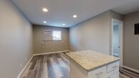 This single-floor space showcases a clean, contemporary design with consistent wood-look luxury vinyl flooring running throughout. The kitchen serves as a central highlight, featuring crisp white shaker-style cabinetry, granite countertops with warm speckled tones, and a full suite of stainless steel appliances including a range, over-the-range microwave, dishwasher, and refrigerator. A white subway tile backsplash adds a polished finishing touch. A peninsula with white cabinetry and a granite countertop extends into the adjacent living area, providing additional workspace or casual seating. Recessed lighting illuminates the open layout effectively, complemented by neutral gray-toned walls that create a cohesive, modern palette throughout. A window-mounted air conditioning unit provides climate control in the main living area. The bathroom is well-appointed with a full tub-shower combination featuring a clean white surround, a white vanity, and matching flooring. A closet with double doors provides practical storage. The space benefits from a private entrance door, adding a sense of independence and convenience to the overall layout.