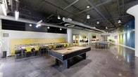 This single-floor recreational space offers an open, industrial-inspired environment designed for both leisure and social interaction. The exposed ceiling reveals structural beams, ductwork, and conduit, all finished in dark tones that complement the polished concrete floors below, creating a raw yet intentional aesthetic throughout.

At the center of the space sits a full-size billiards table, serving as the focal point of the room. To one side, a casual dining and lounge area features sleek yellow chairs paired with light wood tables, set against a decorative tiled accent wall with warm yellow paneling above — adding a pop of color and energy to the otherwise neutral palette.

Further into the space, ping pong tables provide additional recreational opportunities, making the area well-suited for group activities and social gatherings. The layout flows openly between zones, allowing easy movement and visibility across the entire floor.

Overhead, a combination of industrial pendant lights and track lighting ensures the space is well-lit while reinforcing the contemporary, urban character of the design. Beyond the main recreational area, doorways lead to additional rooms visible in the background, suggesting further amenities and flexible-use spaces. Overall, the space strikes a balance between functional design and an energetic, welcoming atmosphere.