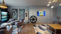 This single-floor communal space showcases a thoughtfully curated interior that merges industrial character with modern design sensibilities. The layout is divided into distinct zones that flow naturally into one another, unified by a cohesive cool-gray color palette and dark-trimmed doors that add sharp contrast throughout.

The primary lounge area features rich, wide-plank hardwood flooring and a large sectional leather sofa anchored beneath subtle overhead lighting. Bold abstract artwork adorns the gray walls, contributing an artistic edge to the relaxed atmosphere. An adjacent seating nook transitions to lighter hardwood flooring and is anchored by geometric-patterned area rugs in blue and neutral tones. This zone incorporates upholstered armchairs, a wall-mounted flat-screen television, and a striking large industrial gear sculpture set against a corrugated metal accent wall—a defining focal point of the space.

A built-in coffee and refreshment station with navy cabinetry and corrugated panel detailing adds functional charm. Natural light enters through multiple windows offering outdoor views, complemented by a pendant light fixture in a warm metallic finish. The overall ambiance is modern, eclectic, and inviting, making this space well-suited for community gathering and relaxation.