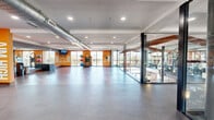 This single-floor fitness center is thoughtfully designed to accommodate a full range of workout activities. The space is divided into distinct functional zones, each outfitted to support specific training needs. The open group exercise studio features a polished concrete floor, exposed industrial-style ductwork along the ceiling, and floor-to-ceiling windows that flood the area with natural light and outward views. Bold orange accent walls with motivational graphics — including "Focus," "Feel the Burn," and "Just Do It" — energize the environment throughout. A glass partition wall separates the studio from the strength and cardio areas, maintaining visual connectivity across the facility. The strength training zone is equipped with cable machines, multi-use weight benches, a squat rack, and a comprehensive dumbbell rack, complemented by large wall-mounted mirrors that amplify the sense of space. The cardio zone lines the window-facing perimeter and features a row of treadmills and elliptical machines, allowing users to exercise with panoramic outdoor views. Recessed lighting provides consistent illumination throughout, while rubber flooring in the equipment areas ensures safety and durability. The overall design balances industrial aesthetics with modern fitness functionality.