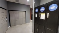 This single-floor amenity space is designed to streamline everyday conveniences for its users, combining package management and laundry services in a clean, functional environment. The space features a bank of gray parcel lockers along one wall, offering secure individual compartments for package storage and retrieval. Complementing these are sleek black Laundry Doctor drop-off lockers, branded with the service's recognizable blue logo, which provide a simple three-step process — drop, lock, and go — making laundry pickup and delivery effortless. The corridor-style layout is finished with light polished flooring and smooth gray walls, creating a bright and modern aesthetic. Recessed ceiling lighting ensures the area is well-illuminated throughout. Unit doors are clearly numbered along the hallway, adding to the organized and orderly feel of the space. The overall design prioritizes efficiency and ease of use, making it a practical hub for daily resident needs.