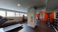 This fitness center is thoughtfully designed to support a full range of workout routines within a single, open floor plan. The space is divided into two functional zones: a cardio area and a strength training area, separated by a structural column that serves as a design focal point, featuring a bold motivational quote reading "Don't stop when you are tired, stop when you are done."

The cardio zone is lined with windows along one wall, allowing natural light to filter in while providing views of the surrounding greenery. Equipment includes multiple treadmills, an elliptical trainer, and a stationary bike, all arranged on durable rubber flooring. A wall-mounted television adds convenience for users during longer cardio sessions.

The strength training zone transitions with a vibrant orange accent wall that energizes the space. It houses a multi-station cable machine, adjustable weight bench, and free weights, accompanied by a large wall mirror for form monitoring. A graphic silhouette of a weightlifter with the phrase "Impossible is nothing" reinforces the motivational atmosphere.

Recessed lighting throughout ensures consistent illumination, and a mini-split HVAC unit maintains climate comfort. An attached restroom with tile flooring, pedestal sink, and vanity lighting adds practical convenience to the facility.