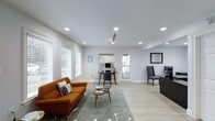 This well-appointed single-floor space offers a clean, modern aesthetic defined by a neutral color palette, light hardwood-style flooring, and a smooth white ceiling equipped with recessed lighting and a sleek track light fixture. The open layout seamlessly integrates a comfortable lounge area, anchored by a warm burnt-orange sofa, a round coffee table, and a soft area rug, creating an inviting space for relaxation or informal gatherings. Multiple workstations are thoughtfully positioned throughout the room — one featuring a desktop computer setup with a mid-century style desk and chair, and another with a dark executive desk and upholstered chair — making the space highly functional for productivity. Large windows fitted with white blinds line the walls, allowing generous amounts of natural light to fill the interior and enhance the airy atmosphere. A glass-paneled door at the rear of the space provides access to an outdoor area, further contributing to the bright and open feel. The overall ambiance is polished yet approachable, balancing modern design elements with practical furnishings suited for both work and leisure.
