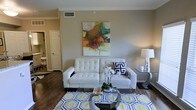 This single-story Dallas residence showcases a thoughtfully designed living space that blends modern aesthetics with comfortable functionality. The living area serves as the centerpiece, featuring rich wood-look flooring that anchors the room with warmth and visual depth. Neutral, warm-toned walls provide a versatile backdrop, complemented by a bold abstract canvas painting that introduces vibrant pops of color and artistic character to the space.

The room is furnished with a tufted white leather sofa, offering a clean, contemporary focal point, paired with a round glass-top coffee table that adds a light, open feel. A geometric-patterned area rug in black, white, and yellow defines the seating area with graphic visual interest. Accent pieces, including a modern table lamp and fresh floral arrangement, contribute to the polished, curated ambiance.

Doorways visible from the living area suggest a practical, connected layout, with access to additional rooms including what appears to be a bathroom. Ceiling-mounted HVAC vents indicate a climate-controlled environment designed for year-round comfort. Overall, the space presents a modern, well-appointed interior suited for stylish everyday living.
