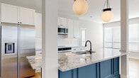 Located at 1140 Chambers Rd in Columbus, Ohio, this thoughtfully designed 2-bedroom, 2-bathroom residence offers a refined living experience across a single, well-appointed floor. The open-concept kitchen and living area serves as the heart of the home, showcasing a striking blue kitchen island topped with elegant granite countertops, white shaker cabinetry, a decorative arabesque tile backsplash, and stainless steel appliances. Globe pendant lights add a sophisticated finishing touch overhead, while light hardwood flooring flows throughout the main living areas, enhancing the bright, airy atmosphere. The primary bedroom is generously sized, featuring plush carpet, multiple windows for ample natural light, and an accent wall adorned with classic raised panel molding. It connects to a well-appointed bathroom complete with marble-look tile flooring, a glass-enclosed shower, and built-in shelving. The second bedroom also benefits from hardwood flooring and large windows. Two closets provide practical storage solutions, with built-in shelving and hanging rods. Crisp white panel doors with matte black hardware carry a consistent modern aesthetic throughout the hallways and rooms, tying together a clean, contemporary design sensibility.
