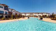 This single-floor residence in Richmond, TX, 77407, is part of a modern multi-building community designed with contemporary aesthetics and upscale amenities. The community's centerpiece is a sweeping resort-style pool with a freeform shape, surrounded by lounge chairs, shade umbrellas, and lush landscaping set against clear Texas skies.

Inside, the open-concept layout connects the kitchen, dining, and living areas seamlessly. The kitchen is outfitted with granite countertops, gray shaker-style cabinetry, stainless steel appliances, and a large kitchen island with an undermount sink, complemented by striking pendant lighting overhead. Light wood-style flooring runs throughout, adding warmth and continuity.

The dining area features built-in shelving and flows naturally into the living space, which is bright and airy with windows allowing natural light. The living room includes a bold accent wall and access to an outdoor area via glass doors.

Throughout the unit, crisp white walls, modern fixtures, and thoughtful design details contribute to a clean, contemporary ambiance well-suited to comfortable everyday living in the Richmond area.