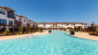 This single-floor residence in Richmond, TX, 77407, is part of a modern multi-building community designed with contemporary aesthetics and upscale amenities. The community's centerpiece is a sweeping resort-style pool with a freeform shape, surrounded by lounge chairs, shade umbrellas, and lush landscaping set against clear Texas skies.

Inside, the open-concept layout connects the kitchen, dining, and living areas seamlessly. The kitchen is outfitted with granite countertops, gray shaker-style cabinetry, stainless steel appliances, and a large kitchen island with an undermount sink, complemented by striking pendant lighting overhead. Light wood-style flooring runs throughout, adding warmth and continuity.

The dining area features built-in shelving and flows naturally into the living space, which is bright and airy with windows allowing natural light. The living room includes a bold accent wall and access to an outdoor area via glass doors.

Throughout the unit, crisp white walls, modern fixtures, and thoughtful design details contribute to a clean, contemporary ambiance well-suited to comfortable everyday living in the Richmond area.