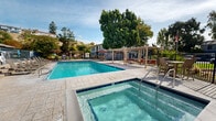 Located at 813 Southampton Rd in Benicia, California, this two-story property offers a well-appointed outdoor amenity area that serves as a central gathering space for residents. The outdoor area is anchored by a generously sized swimming pool with crystal-clear blue water, complemented by a dedicated spa that provides a relaxing retreat. The pool deck is finished with attractive paved stonework and is furnished with multiple lounge chairs, dining sets, and large shade umbrellas, creating comfortable spaces for both lounging and outdoor dining. A pergola-covered seating area adds architectural interest and provides additional shaded space for socializing. A barbecue grill station is also available, making the outdoor area well-suited for gatherings. The property is enclosed by a wrought iron fence and is framed by mature deciduous trees that provide natural shade and seasonal color. The surrounding hillside landscape and open California sky contribute to a bright and airy ambiance throughout the outdoor space. The two-story building adjacent to the pool area features a classic residential exterior with a neutral color palette, providing a backdrop that blends seamlessly with the natural surroundings. Overall, the property combines functional outdoor amenities with a relaxed, park-like setting in the heart of Benicia.