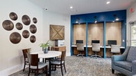 This well-appointed business center offers a thoughtfully designed workspace that balances productivity and comfort. The centerpiece of the room is a striking accent wall finished in bold blue, housing three individual built-in workstations crafted from warm natural wood. Each station is equipped with a monitor and illuminated by dedicated adjustable wall-mounted sconce lighting, creating a focused and functional environment for individual work.

The open layout also features a round white dining-height table with dark wood chairs, ideal for collaborative meetings or casual gatherings. Comfortable upholstered accent chairs are positioned throughout, providing flexible seating for lounging or informal conversations. A wooden sideboard along one wall accommodates a printer and office essentials, adding practical utility to the space.

The room is decorated with curated wall art, including circular metallic disc arrangements, a geometric wood-inlay panel, and framed artwork, lending the space a warm, contemporary aesthetic. Patterned carpet in earthy tones anchors the room and adds visual texture underfoot.

Large floor-to-ceiling windows and a glass exterior door flood the space with natural light and offer views of the surrounding outdoor areas, including a patio with seating and lush greenery. Glass-paneled interior doors connect the business center to adjacent spaces, enhancing the sense of openness and connectivity throughout.