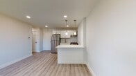 This thoughtfully designed single-floor unit offers a clean, modern aesthetic throughout, with a cohesive palette of soft whites, light grays, and warm wood-tone luxury vinyl plank flooring that flows through the main living areas. The open-concept kitchen anchors the space, showcasing white shaker-style cabinetry with sleek bar-pull hardware, bright white countertops, and a striking herringbone-patterned tile backsplash. Stainless steel appliances — including a gas range and full-size refrigerator — complete the kitchen, while a peninsula with pendant lighting above provides additional prep space and a casual dining or seating option. The living area is open and well-proportioned, bathed in recessed lighting and offering a comfortable, versatile layout. The bedroom features plush wall-to-wall carpeting, a stylish sputnik-style chandelier, and a window that lets in natural light with pleasant greenery views. The full bathroom is neatly finished with white subway tile surrounding the tub-shower combo, a decorative mosaic accent border, dark-toned floor tile, and a white vanity with ample storage. A standout convenience is the dedicated in-unit laundry area, where a stacked washer and dryer are neatly tucked into an alcove, adding everyday practicality to this well-appointed space.