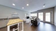 This single-floor space showcases a thoughtfully designed open-concept layout that integrates the kitchen and living areas into one cohesive, functional environment. The interior is finished with warm hardwood-style flooring that runs throughout, complementing the neutral grey walls and textured ceiling to create a clean, contemporary aesthetic.

The kitchen area features open white shelving mounted along the wall, providing accessible storage and display space for everyday essentials. A butcher block-topped island or counter adds a natural wood element, offering additional workspace and a visual contrast to the otherwise cool-toned palette. Recessed lighting is installed throughout the ceiling, ensuring consistent, well-distributed illumination across the space.

The living area is generously sized and anchored by a low-profile media console supporting a flat-screen television. A plush sectional sofa, accent chairs, a glass-top coffee table, and a cowhide-style area rug come together to define a comfortable and stylish seating arrangement. Large windows along the far wall invite abundant natural light and offer views of the surrounding greenery, while a glass door provides direct access to the outdoors, enhancing the sense of openness and connectivity to the exterior.