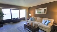 This single-floor space centers around a thoughtfully designed living room that balances comfort and style. The room features a striking contrast between a deep chocolate-brown accent wall and lighter neutral tones on the surrounding walls and carpet, creating a warm, inviting atmosphere. A large picture window serves as a focal point, flooding the space with natural light while offering expansive views of the surrounding cityscape and waterfront beyond. The carpeted floor adds to the cozy feel underfoot. Furnishings include a generously sized cream-toned sectional sofa accented with blue and patterned throw pillows, positioned around a dark wood coffee table with drawer storage. A patterned area rug in blue and white anchors the seating arrangement. On the accent wall, a recessed display shelf with a framed mirror adds architectural interest and depth. A small side table with a lamp provides warm ambient lighting. Decorative touches such as ceramic vases, potted greenery along the windowsill, and subtle wall script reading "Smile" and "Breathe" contribute to the space's relaxed, welcoming character.