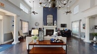 This single-story clubhouse in Horn Lake, Mississippi, makes a strong architectural impression through its dramatically tall ceilings, which lend an open, airy quality throughout the entire space. Dark, richly toned tile flooring anchors the interior, contrasting beautifully with crisp white walls and decorative crown molding.

The main gathering area serves as the centerpiece of the clubhouse, highlighted by a striking navy blue accent wall that frames a classic white fireplace mantle. A large arched mirror mounted above the mantle adds depth and elegance, while ceiling fans and a statement chandelier overhead provide both function and visual interest. The lounge area is furnished with comfortable seating arrangements, coffee tables, and area rugs in coordinating blue and neutral tones, creating an inviting atmosphere for residents to gather.

Adjacent to the lounge is a dining area with dark table and chair sets, accompanied by a television mounted on the wall. A dedicated workspace with computer stations and desks provides a practical area for business needs or remote work.

The leasing office area, accessible near the entrance, features a large dark wood desk, decorative columns, and French-style glass doors that allow natural light to flow freely inward. Multiple windows throughout the clubhouse further enhance the bright, welcoming ambiance of this well-appointed community facility.