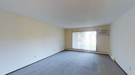 This single-floor space features a spacious, open living area finished with wall-to-wall carpet in a neutral grey tone, contributing to a clean and understated aesthetic. The room is characterized by smooth, white ceilings and light beige walls that create a bright, airy atmosphere throughout. A large picture window serves as the focal point of the main living area, fitted with vertical blinds that allow for flexible light control while offering a view of the surrounding greenery outdoors. Natural light filters generously through the window, enhancing the open feel of the room. A wall-mounted air conditioning unit is positioned adjacent to the window, providing in-unit climate control. Baseboard heating runs along the base of the exterior wall, offering an additional source of warmth. The layout is open and adaptable, providing ample room for a variety of furniture arrangements. Electrical outlets are conveniently placed along the walls. Overall, the space presents a functional and practical interior with a simple, neutral palette well-suited to a range of décor styles.