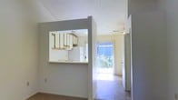 This single-floor residence offers a straightforward, functional layout designed to maximize connectivity between its living spaces. The kitchen is defined by a pass-through window cut into the dividing wall, creating an open sightline into the main living area. Upper wood-trimmed cabinetry lines the kitchen walls, providing ample storage, while a white refrigerator and a sink with a light countertop complete the functional workspace. The textured ceiling adds a subtle architectural detail throughout the space.

The living area flows naturally toward the back of the residence, where a full-height sliding glass door floods the space with natural light and provides direct access to an outdoor balcony or patio. Vertical blinds frame the door, offering privacy and light control. The flooring transitions between carpet in the living zone and tile near the entryway and sliding door, offering both comfort and practicality. A ceiling fan is mounted overhead in the living area, contributing to climate control. Neutral gray and white walls throughout maintain a clean, cohesive aesthetic. A secondary doorway is visible along the side wall, suggesting access to additional rooms beyond the main living space.