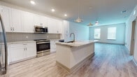 This single-floor unit offers a clean, contemporary design throughout, unified by light-toned luxury vinyl plank flooring that flows seamlessly from room to room. The centerpiece of the space is the open-concept kitchen, which is outfitted with crisp white shaker-style cabinetry, a neutral tile backsplash, and granite countertops. A generously sized island with an undermount sink and gold-toned faucet provides additional prep space and serves as a natural gathering point. Stainless steel appliances, including a French door refrigerator, gas range, built-in microwave, and dishwasher, complete the kitchen's modern, functional aesthetic. Stylish pendant lights hang above the island, complementing the recessed lighting overhead. The kitchen opens directly into a bright living area illuminated by natural light from a window and fitted with a ceiling fan. The bedrooms are well-proportioned, each featuring ceiling fans, large windows with blinds, and light-filled interiors. One bedroom includes access to a walk-in closet with built-in shelving. The bathroom presents a spa-like atmosphere with large-format floor tiles, a frameless glass sliding shower enclosure with full tile surround, a white vanity with granite countertops, and chrome fixtures throughout.