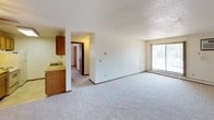 This single-floor unit offers a practical and straightforward layout connecting several distinct functional areas. The galley-style kitchen is efficiently designed with wood-toned cabinetry lining both walls, providing ample upper and lower storage. Light-colored laminate countertops run the length of both sides, with a double stainless steel sink on one end and a white electric coil-burner range with a refrigerator on the other. Overhead fluorescent lighting illuminates the tile floor throughout the kitchen.

The living area is generously proportioned, featuring neutral wall-to-wall carpet, a textured popcorn ceiling, and a large sliding glass door fitted with vertical blinds that allows natural light to filter in. A separate bedroom also features carpet, a ceiling fan with light fixture, baseboard heating, and a window with vertical blinds offering an outdoor view. The room's neutral tones create a calm, understated atmosphere.

The bathroom includes a standard tub and shower combination with chrome fixtures and towel bars, a toilet, and a vanity with countertop storage. Tile flooring and white walls give the bathroom a clean, functional appearance. Overall, the unit provides a no-frills, well-organized living environment.
