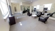 This single-floor professional space presents a well-appointed interior designed for comfort and functionality. The main area serves as a reception or lounge, featuring plush dark leather armchairs arranged around a contemporary round metallic coffee table, creating an inviting atmosphere for visitors and clients alike. Large wall-mounted mirrors span a significant portion of the room, visually expanding the space and amplifying the natural light that streams in through the windows. The neutral-toned carpet throughout provides a clean, polished foundation that complements the crisp white walls and recessed ceiling lighting. A dark wood reception desk anchors one side of the room, reinforcing the professional character of the space. A framed artwork piece adorns one wall, adding a subtle decorative touch without detracting from the overall refined aesthetic. Additional workstations and equipment are visible in the background, suggesting dedicated functional areas beyond the main lounge. The layout is open and uncluttered, allowing for easy movement throughout the space. A private room with a frosted or solid door offers separation for more confidential functions. Overall, the space strikes a balance between a welcoming reception environment and an efficient, modern professional setting.
