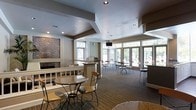 This single-floor communal space is thoughtfully designed to foster comfort and social interaction. The interior is anchored by a striking herringbone-pattern brick fireplace, serving as a warm and elegant focal point that draws the eye upon entry. Recessed lighting is embedded throughout the tray-style ceiling, complementing the natural light that floods in through a generous arrangement of large windows and glass-paneled doors along the far wall, offering views of the greenery outside.

The layout is organized into distinct seating zones, catering to both intimate gatherings and larger groups. Round bistro-style tables with sleek metal chairs occupy the main lounge area, while bar-height tables and stools line the perimeter near the windows, providing a casual yet functional setting. A centrally positioned counter or service station adds a practical element to the space.

Neutral tones in taupe and warm gray are carried throughout the walls and carpeted flooring, creating a cohesive and inviting atmosphere. Subtle architectural details, including stepped ceiling elements and clean structural columns, add visual depth without overwhelming the space. Overall, this lounge exudes a polished yet approachable character, making it a versatile venue for everyday use or organized events.