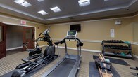Located at 46 River St in the Mattapan neighborhood of Boston, MA, this fitness center offers a dedicated space for exercise and wellness. The room features warm golden-beige walls accented by white chair rail molding and crown molding, lending a clean and polished appearance. Striped carpeting covers the floor throughout, providing a practical surface across the workout area.

The facility is equipped with a diverse selection of exercise machines, including treadmills, elliptical trainers, a stationary bike, and a rowing machine, all arranged to allow comfortable movement between stations. A dedicated free weight area includes a dumbbell rack, a weight bench, and a stability ball, catering to strength training needs. A wall-mounted flat-screen television is positioned for visibility during cardio workouts.

A floor-to-ceiling mirror along one wall enhances the sense of space and allows users to monitor their form. The room is well illuminated by recessed panel lighting in a drop ceiling. A built-in bench provides a seating area near the entry, and a wall-mounted paper towel dispenser and utility sink support hygiene and equipment maintenance. Double wood-and-glass doors provide clear access in and out of the space.
