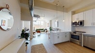 Located at 57 Cannon St in Poughkeepsie, NY, this modern loft-style unit blends industrial character with contemporary finishes across a single open floor plan. The living area is anchored by dramatically high ceilings with exposed metallic ductwork, light hardwood flooring, and oversized windows that draw in generous natural light and offer urban views of the surrounding streetscape. The open-concept kitchen is thoughtfully appointed with crisp white shaker cabinetry, quartz countertops, a waterfall-edge island with bar seating, stainless steel appliances including a range, microwave, refrigerator, and dishwasher, as well as open shelving and globe pendant lighting that adds a warm, modern touch. The living space flows seamlessly from the kitchen, providing ample room for lounging and entertaining. The bedrooms are calm and comfortable retreats, each featuring high ceilings, soft neutral walls, and windows that bring in natural light; one bedroom also includes a dedicated workspace area. The bathroom is finished with striking herringbone-pattern wall tile, a white vanity with matte black fixtures, and wood-look flooring, striking a balance between style and function throughout.