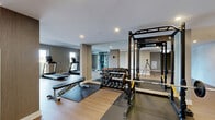 Located at 265 Boulevard Brunswick in Pointe-Claire, Quebec, this well-appointed fitness facility offers a generously sized, single-floor exercise room designed to support a full range of workout routines. The space features light wood-toned flooring throughout, neutral grey and taupe walls, and recessed lighting that creates a bright, clean atmosphere. Structural columns clad in warm wood-finish panels add an architectural dimension to the open layout, while large sliding windows along one wall flood the cardio area with natural light and offer views of the surrounding greenery. The strength training zone is anchored by an Ironax power rack and cable machine system, complemented by a full dumbbell rack and a weight bench set on protective floor mats. A floor-to-ceiling mirror wall visually expands the space and supports form monitoring during workouts. The cardio section accommodates multiple treadmills, stationary spin bikes, and an elliptical trainer, all positioned to take advantage of the natural light from the exterior windows. A wall-mounted television and a dedicated climate control unit ensure comfort and entertainment during exercise. The facility also includes open floor space suitable for stretching, functional training, or group activity. The overall design is modern and functional, balancing practical fitness amenities with a clean, inviting aesthetic.