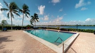 This waterfront property is defined by its exceptional outdoor amenity space, centerpiece of which is a generously sized swimming pool with striking blue-tiled accents and a classic rectangular design. The pool deck, finished in warm-toned brick pavers, provides ample space for lounging and outdoor entertaining. Stainless steel entry rails offer easy pool access, while the surrounding landscape features mature, swaying palm trees that frame the setting with a distinctly tropical character. The property sits directly along a broad waterway, and the unobstructed views across the open water create a serene, resort-like ambiance. Manicured hedgerows define the perimeter of the outdoor space, adding a sense of privacy while preserving the panoramic vistas. Additional seating areas are positioned along the water's edge, ideal for taking in the scenery. The expansive sky above, with its rolling clouds and abundant natural light, enhances the open, airy feel of the outdoor environment. The overall setting reflects a warm, coastal lifestyle with permanent features designed to maximize the enjoyment of the waterfront location and South Florida's year-round outdoor living conditions.