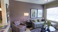 Located in Lewisville, TX, this single-story space offers a thoughtfully designed interior that blends contemporary style with a warm, inviting ambiance. The living area serves as the heart of the space, showcasing a bold plum-toned accent wall that creates a striking visual backdrop. A neutral-toned sofa is dressed with green and patterned accent pillows, complemented by a coordinating tufted armchair in muted purple stripes — together creating a cohesive and sophisticated color palette. A sleek black side table with decorative metallic accents adds a modern touch, while a tall floor lamp with a clean white shade provides warm, functional lighting. Two framed art pieces are displayed prominently on the accent wall, adding a gallery-like quality to the room. A patterned area rug with floral motifs anchors the seating arrangement and ties together the earth-toned and jewel-toned color scheme. The overall aesthetic is polished and curated, reflecting an attention to detail in both furnishing selections and spatial arrangement. The single-floor layout offers ease of movement and accessibility throughout, making it a practical yet visually refined living environment.