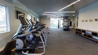 This single-floor fitness center offers a thoughtfully designed workout environment that balances functionality with a clean, modern aesthetic. The space is illuminated by striking linear LED lighting fixtures mounted across the ceiling at dynamic angles, providing bright, even illumination throughout. Large windows along one wall invite generous natural light and offer views of the surrounding greenery, creating an open and energizing atmosphere.

The cardio zone is prominently featured along one side of the room, lined with a row of elliptical trainers and treadmills, offering ample options for aerobic exercise. The flooring throughout is durable, low-pile carpet in a neutral tone, providing both comfort underfoot and sound absorption. Toward the rear of the space, a multi-functional cable and strength training machine system serves as a dedicated zone for resistance workouts, accompanied by a heavy punching bag for boxing or conditioning training. Wall-mounted mirrors near the entrance enhance the sense of openness. The neutral color palette of the walls complements the modern equipment, contributing to a focused yet welcoming fitness environment suitable for a wide range of workout routines.