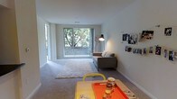 This single-floor space offers a clean, modern aesthetic defined by an open-concept layout that connects the living and dining areas in a natural, functional flow. The living area is anchored by a large window that floods the room with abundant natural light and frames lush tree views beyond a private balcony, creating a refreshing indoor-outdoor connection. Light-toned carpeting and crisp white walls contribute to an airy, welcoming ambiance throughout. The living space comfortably accommodates a sofa and area rug, while a floor lamp adds warm accent lighting. The dining area sits adjacent, featuring a casual table setup that complements the relaxed, contemporary feel of the space. Walls are left largely open, offering a neutral canvas that adapts easily to a variety of décor styles. The overall design prioritizes openness, light, and simplicity, making the space feel both functional and inviting. Sliding access to the balcony further enhances the sense of space, connecting the interior to the outdoors and allowing for excellent natural ventilation and light throughout the day.