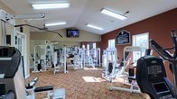 Located at 3851 Cape Landing Drive in Myrtle Beach, South Carolina, this fitness center offers a comprehensive and well-maintained workout facility designed to meet a variety of exercise needs. The single-floor layout is open and spacious, featuring vaulted ceilings that enhance the sense of airiness throughout the room. Large windows allow natural light to filter in, complementing the bright overhead fluorescent lighting that keeps the space well-illuminated at all times. The facility is outfitted with an extensive selection of strength training machines, including cable systems, press machines, and various resistance equipment, all arranged to allow comfortable movement between stations. Full-length mirrors line one wall, providing users with visual feedback during workouts and further amplifying the sense of space. A wall-mounted flat-screen television is positioned for easy viewing from multiple areas of the floor. The accent wall, finished in a deep terracotta red, adds a bold visual contrast to the neutral tones of the surrounding walls and ceiling. The patterned carpeted flooring provides both comfort and durability underfoot. A posted information board on the accent wall offers facility rules and guidelines for members. Overall, the space delivers a functional, energizing environment suited for a full strength-training regimen.