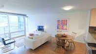 This well-appointed single-floor residence offers an open-concept layout that combines the living and dining areas into one cohesive, light-filled space. Warm hardwood flooring runs throughout, providing a grounding contrast to the crisp white walls and ceiling. Large floor-to-ceiling windows flood the interior with natural light while offering views of the surrounding cityscape, enhancing the sense of openness and airiness.

The living area is anchored by a clean-lined white sofa, complemented by a low media console housing a flat-screen television. A wall-mounted piece of vibrant pop-art — featuring bold florals in red, orange, and yellow — adds a striking focal point and a splash of color to the otherwise neutral palette.

The dining area features a rustic round wooden table paired with matching wooden chairs, creating a warm and inviting spot for meals. A bowl of fresh fruit on the table adds a homely touch. The combination of modern and natural elements throughout gives the space a balanced, welcoming ambiance that is both functional and aesthetically refined.