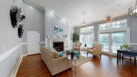This single-floor space offers an inviting and well-appointed layout designed for both comfort and functionality. The main lounge area immediately draws attention with its soaring vaulted ceilings, elegant wainscoting panels, and soft gray walls that create a calm, sophisticated atmosphere. Rich hardwood flooring flows throughout the entire space, adding warmth and continuity. A prominent stone-surround fireplace serves as the focal point of the lounge, topped with a crisp white mantel and framed by decorative paneling that extends to the ceiling. Thoughtfully arranged seating, including a plush neutral sofa and upholstered accent chairs, encourages relaxation and conversation. Large windows with transom panels flood the space with abundant natural light and provide views of the surrounding greenery, while French doors offer direct access to the outdoors. A geometric pendant chandelier adds a refined touch overhead. Decorative wall art and lush potted plants enhance the overall aesthetic. The kitchen features white cabinetry, granite-style countertops, and a central island with an integrated sink, pendant lighting, and ample prep space. An adjacent dining area with decorative arched windows completes the layout, making this a versatile and welcoming communal environment.