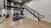 This fitness facility is designed to accommodate a comprehensive workout experience across a thoughtfully arranged split-level layout. The main floor offers generous open space with light wood flooring and protective rubber mats, providing ample room for free weight training, stretching, and functional fitness exercises. A dumbbell rack, adjustable bench, and cable machine are among the equipment stations positioned throughout the lower level, complemented by a stability ball and additional strength training apparatus.

Soaring ceiling heights lend the space an open, airy feel, while the exposed cinder block walls give the interior an industrial character. A striking staircase with a black metal cable railing leads to an upper mezzanine level, where additional cardio and strength equipment can be found, maximizing the facility's overall training capacity.

The entrance features a full-glass door framed in black, allowing natural light to flow into the reception area, which is equipped with a water cooler and informational signage. A large wall-mounted mirror along the main training floor enhances the sense of space and allows users to monitor their form. The combination of functional design, durable materials, and well-organized equipment makes this a versatile and practical space suited for a wide range of fitness activities.