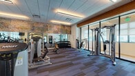 This modern fitness center is thoughtfully designed to accommodate a full range of workout routines across its single-floor layout. The space is defined by a striking exposed brick accent wall adorned with colorful lettering, which adds an industrial-chic character that contrasts beautifully with the clean white walls and drop-tile ceiling fitted with recessed linear lighting. Multi-toned striped carpet tiles in shades of blue, gray, and brown cover the floor throughout, providing both comfort and visual structure to the space.

The main workout area is generously outfitted with Hoist and Cybex branded strength machines, a full dumbbell rack with adjustable benches, cable machines, and multi-functional cable systems. A dedicated cardio zone houses a row of Cybex treadmills and additional cardio equipment, accompanied by wall-mounted flat-screen TVs for entertainment during workouts. Floor-to-ceiling mirrors along one wall amplify the sense of space and allow users to monitor their form.

A glass-partitioned section near the entrance offers an additional workout zone with natural light filtering in through large windows. Near the entry, hand sanitizer dispensers and waste bins are conveniently placed. The overall ambiance is clean, professional, and energizing — a well-rounded facility suited for both strength training and cardiovascular exercise.